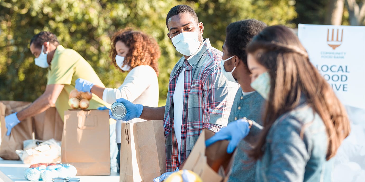 A mid adult man places a canned food item into a paper bag as he volunteers with his teenage son at an outdoor food bank. He and other volunteers are wearing protective face masks as they are volunteering during the coronavirus pandemic.