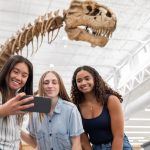 Three young women take a selfie in front of a big dinosaur skeleton in a library lobby.